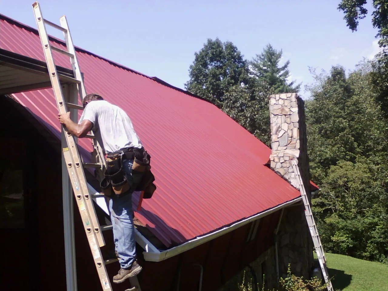 Roofer on a ladder beside a red metal roof and stone chimney — Soffit & Fascia Repair by Roofing Brothers in Waveland